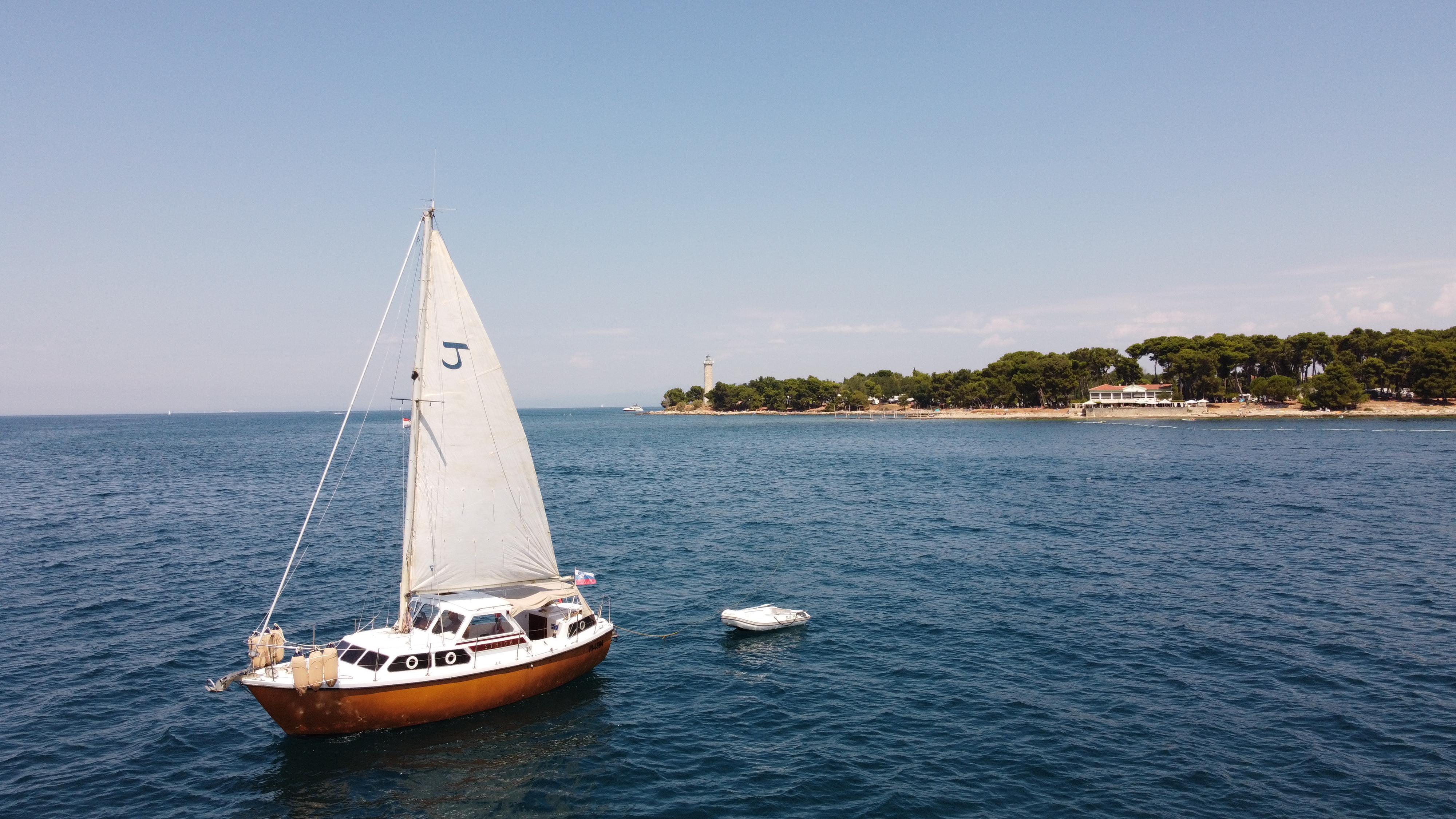Aerial drone view of STRIGA under sail with her tender in tow, approaching the Piran coastline and lighthouse